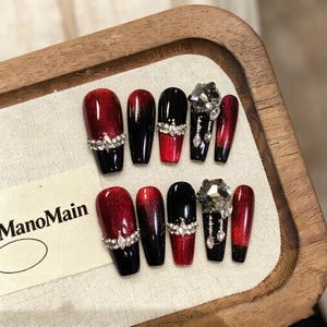 Puede incluir: Un conjunto de uñas postizas en una bandeja de madera. Las uñas presentan un diseño degradado de rojo a negro, algunas adornadas con pedrería y una gema grande. La marca "ManoMain" es visible.