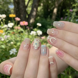 Puede incluir: Primer plano de una mano con manicura que presenta varios diseños de arte de uñas. Las uñas muestran una gama de colores, incluyendo rosa, blanco y azul, con patrones florales y geométricos. Algunas uñas tienen un diseño de flor en 3D y una media luna.
