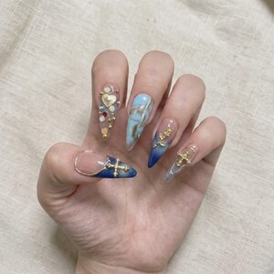May include: Stiletto-shaped artificial nails featuring a variety of designs. One nail has a gold heart and colorful rhinestones, another has blue marble swirls, and others feature blue gradient tips with gold cross and rose metal charms.