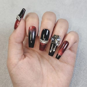 Puede incluir: Uñas postizas largas en forma de ataúd con un degradado de negro intenso y rojo brillante. El diseño incluye adornos de pedrería transparente, con una uña que presenta un gran cristal central rodeado de pequeñas piedras colgantes.
