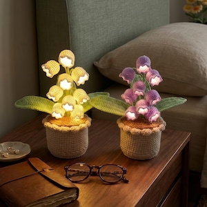 May include: Two crochet flower lamps in woven pots sit on a wooden nightstand. One lamp is yellow, the other purple. Both have green leaves and are lit. A brown journal, glasses, and a small dish with jewelry are also on the table.