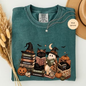 May include: A blue spruce Comfort Colors t-shirt featuring a graphic of two black cats and a ghost wearing witch hats, reading books stacked with titles like Exorcising Fascism, Witches Against Fascism, Saving Democracy, Human Rights, and Civic Duty.