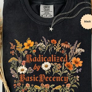 May include: A black crew neck t-shirt featuring a graphic print of various wildflowers in shades of orange, yellow, white, and purple. The text "Radicalized by Basic Decency" is printed in an orange serif font in the centre of the floral design.