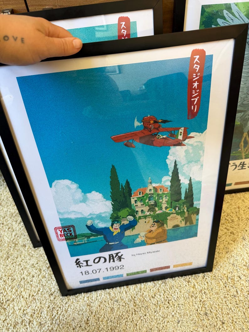 Puede incluir: Impresi&oacute;n art&iacute;stica enmarcada con una vibrante ilustraci&oacute;n de un hidroavi&oacute;n rojo volando en un cielo azul. La obra de arte incluye texto japon&eacute;s y una fecha, con una representaci&oacute;n detallada de un edificio y personajes. La impresi&oacute;n est&aacute; en un marco negro.
