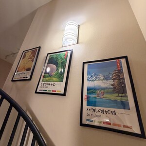 Pu&ograve; includere: Tre poster incorniciati con arte anime giapponese sono appesi a una parete beige, accanto a un'applique cilindrica bianca. I poster presentano illustrazioni colorate e testo giapponese. Una ringhiera in metallo nero &egrave; visibile in primo piano.