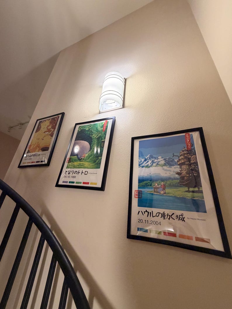 Pu&ograve; includere: Tre poster incorniciati con arte anime giapponese sono appesi a una parete beige, accanto a un'applique cilindrica bianca. I poster presentano illustrazioni colorate e testo giapponese. Una ringhiera in metallo nero &egrave; visibile in primo piano.