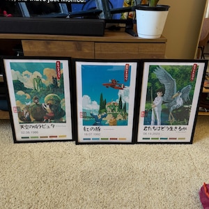 May include: Three framed art prints with black frames. The prints feature colourful illustrations with Japanese text. The first print has a scene with a robot and children. The second print shows a red plane flying over a landscape. The third print depicts a person with a large bird.