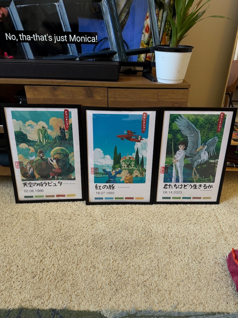 May include: Three framed art prints with black frames. The prints feature colourful illustrations with Japanese text. The first print has a scene with a robot and children. The second print shows a red plane flying over a landscape. The third print depicts a person with a large bird.