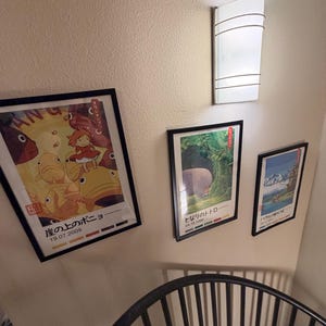 Pu&ograve; includere: Tre poster incorniciati con cornici nere sono montati su una parete beige. I poster presentano arte di animazione giapponese. Un'applique bianca &egrave; montata sopra i poster. Una sedia in metallo nero &egrave; in primo piano.