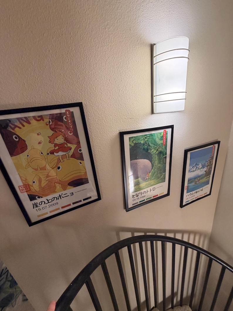 Pu&ograve; includere: Tre poster incorniciati con cornici nere sono montati su una parete beige. I poster presentano arte di animazione giapponese. Un'applique bianca &egrave; montata sopra i poster. Una sedia in metallo nero &egrave; in primo piano.