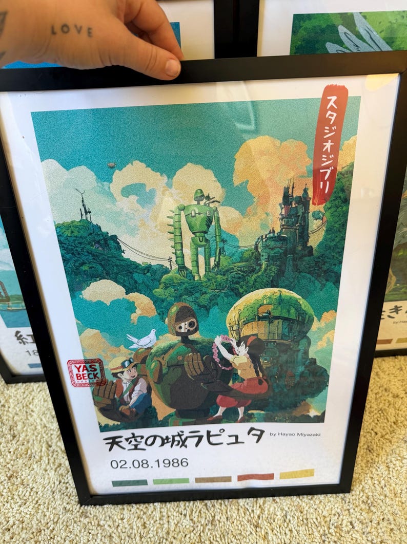 May include: Framed art print with a colourful illustration from "Castle in the Sky." The image shows a green robot, characters, and a floating island. Japanese text and the date 02.08.1986 are also visible in the artwork.