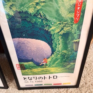 Pu&ograve; includere: Poster incorniciato con una scena del film d'animazione "Il mio vicino Totoro". L'opera raffigura una grande creatura soffice in una foresta con un bambino. Testo giapponese e la data 04.16.1988 visibili.