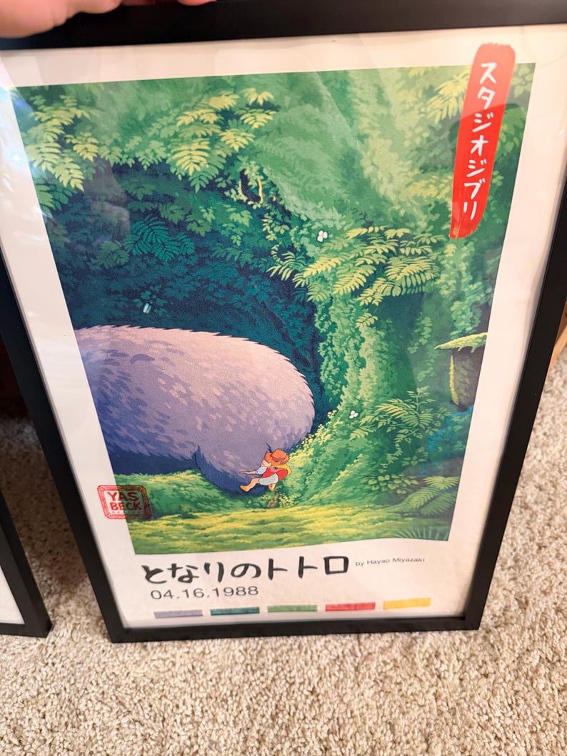 Pu&ograve; includere: Poster incorniciato con una scena del film d'animazione "Il mio vicino Totoro". L'opera raffigura una grande creatura soffice in una foresta con un bambino. Testo giapponese e la data 04.16.1988 visibili.