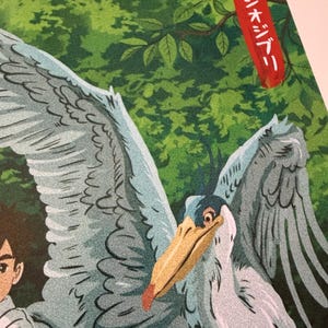 K&ouml;nnte beinhalten: Illustration eines gro&szlig;en Vogels mit ausgebreiteten Fl&uuml;geln, in Graut&ouml;nen, Blau und Beige. Der Vogel hat einen langen Schnabel und ist vor einem gr&uuml;nen Hintergrund aus Bl&auml;ttern platziert. Das Bild enth&auml;lt japanischen Text in einem roten Rechteck.