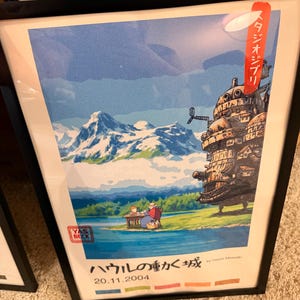 Op de afbeelding: Ingelijste poster van "Howl's Moving Castle" met een bergketen, meer en het kasteel. Japanse tekst en de datum 20.11.2004 zijn zichtbaar. De poster zit in een zwarte lijst.