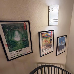 Puede incluir: Tres carteles de anime japon&eacute;s enmarcados cuelgan de una pared beige, con una l&aacute;mpara blanca encima. Los carteles presentan ilustraciones coloridas y texto japon&eacute;s. El cartel inferior tiene la fecha 12.19.1997.