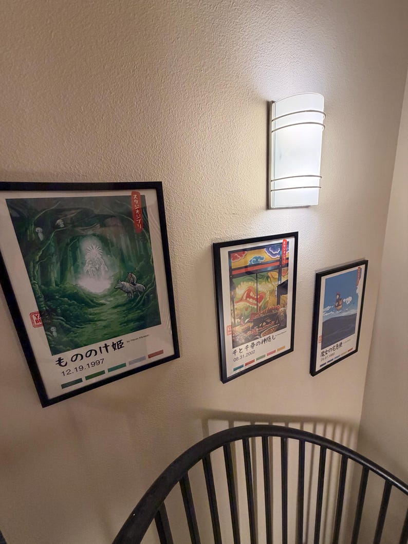 Puede incluir: Tres carteles de anime japon&eacute;s enmarcados cuelgan de una pared beige, con una l&aacute;mpara blanca encima. Los carteles presentan ilustraciones coloridas y texto japon&eacute;s. El cartel inferior tiene la fecha 12.19.1997.