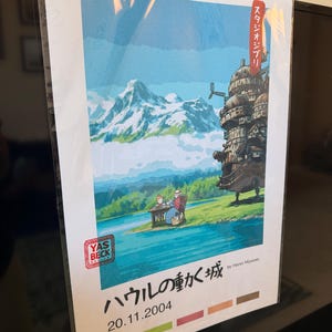 Op de afbeelding: Een ingelijste kunstdruk met een sc&egrave;ne met een meer, bergen en een fantastisch bewegend kasteel. De print bevat Japanse tekst en de datum 20.11.2004. Het kunstwerk wordt op zijn plaats gehouden met een zwarte klem.