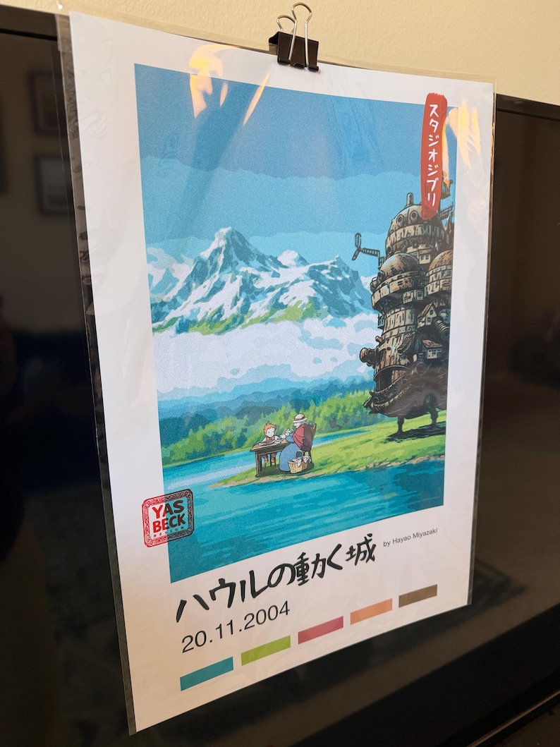 Op de afbeelding: Een ingelijste kunstdruk met een sc&egrave;ne met een meer, bergen en een fantastisch bewegend kasteel. De print bevat Japanse tekst en de datum 20.11.2004. Het kunstwerk wordt op zijn plaats gehouden met een zwarte klem.