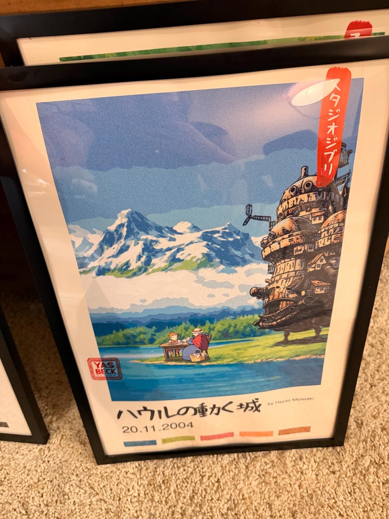 Pu&ograve; includere: Poster incorniciato con una scena de "Il castello errante di Howl". L'opera raffigura un castello, montagne e un lago. Il poster include testo giapponese e la data 20.11.2004. Il poster &egrave; in una cornice nera.