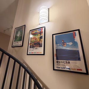 Puede incluir: Tres impresiones art&iacute;sticas enmarcadas con texto japon&eacute;s cuelgan de una pared beige, junto a un aplique de pared cil&iacute;ndrico blanco. Las impresiones presentan ilustraciones coloridas y la barandilla de la escalera es negra.