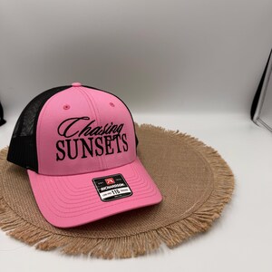 May include: A pink and black trucker hat featuring a mesh back and a solid pink front panel. The text "Chasing SUNSETS" is embroidered in black on the front. A rectangular Richardson 115 brand sticker is affixed to the center of the pink brim.