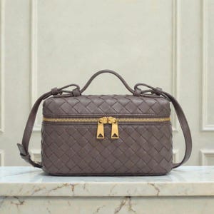 May include: A woven, taupe-colored handbag with a top handle and a gold-toned zipper. The bag has a rectangular shape with a structured design and a crossbody strap. The bag is sitting on a marble surface.