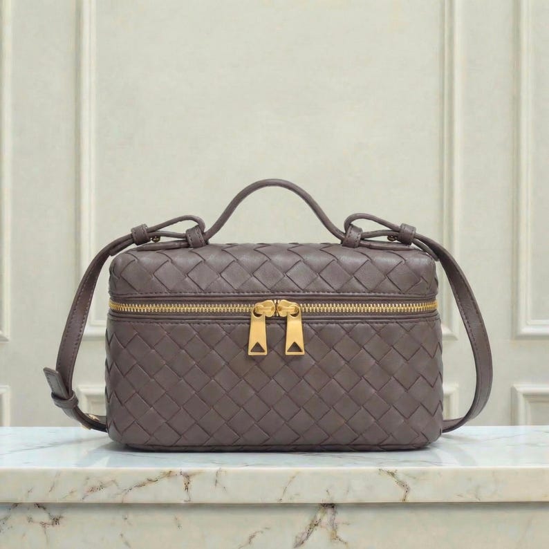 May include: A woven, taupe-colored handbag with a top handle and a gold-toned zipper. The bag has a rectangular shape with a structured design and a crossbody strap. The bag is sitting on a marble surface.