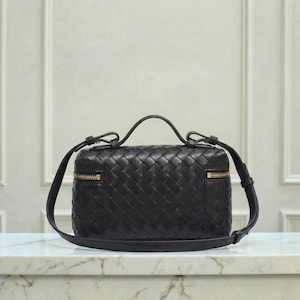 May include: A black leather handbag with a woven pattern. The bag has a top handle and a detachable shoulder strap. Gold-colored zippers are visible on the sides. The bag is rectangular and sits on a marble surface.