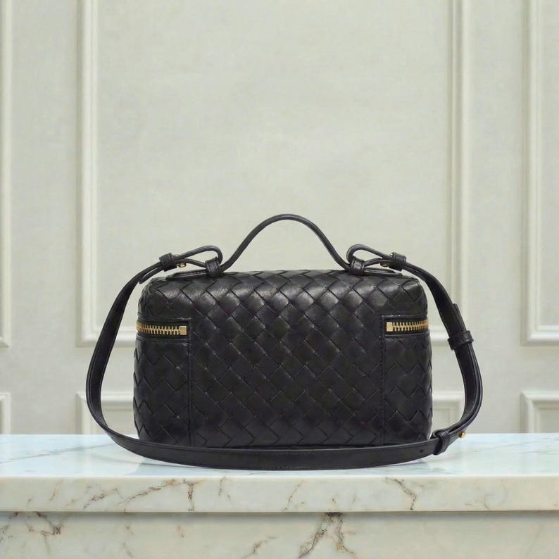 May include: A black leather handbag with a woven pattern. The bag has a top handle and a detachable shoulder strap. Gold-colored zippers are visible on the sides. The bag is rectangular and sits on a marble surface.