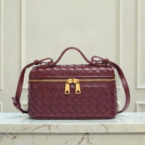 May include: A burgundy leather handbag with a woven design. The bag has a rectangular shape with a top handle and a long, adjustable shoulder strap. It features a gold-tone zipper closure and hardware.