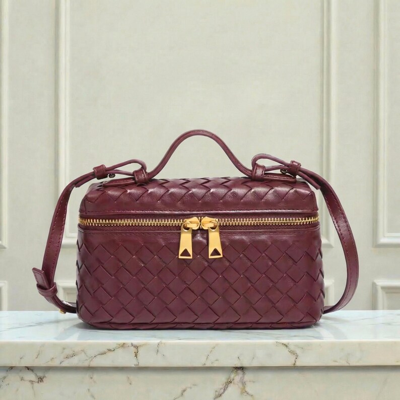May include: A burgundy leather handbag with a woven design. The bag has a rectangular shape with a top handle and a long, adjustable shoulder strap. It features a gold-tone zipper closure and hardware.