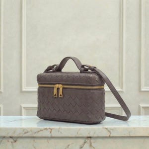 May include: A gray leather handbag with a woven pattern. The bag has a top handle, a long shoulder strap, and a gold zipper. The bag is rectangular and sits on a marble surface.