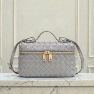May include: A gray, woven leather handbag with a top handle and a gold zipper closure. The bag has a rectangular shape and a crossbody strap. The bag is sitting on a marble surface.
