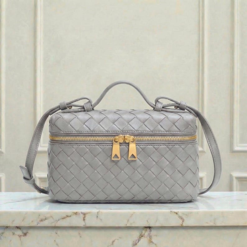 May include: A gray, woven leather handbag with a top handle and a gold zipper closure. The bag has a rectangular shape and a crossbody strap. The bag is sitting on a marble surface.