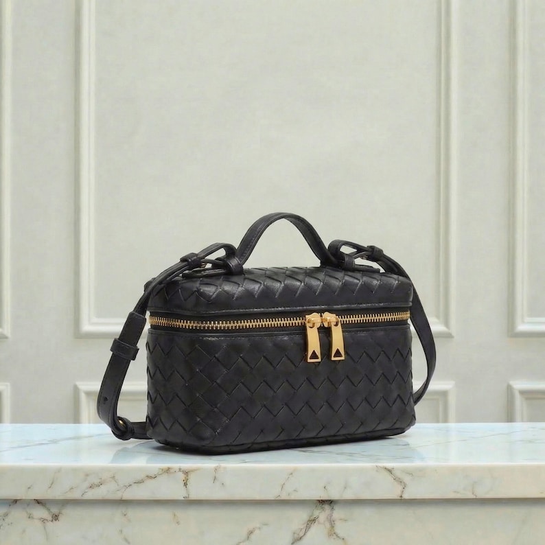 May include: Black leather crossbody bag with a woven design. The bag has a rectangular shape with a top handle and a long adjustable strap. It features a gold-tone zipper and hardware.