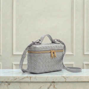 May include: A gray, woven leather crossbody bag with a top handle and a gold-toned zipper. The bag has a rectangular shape and a detachable shoulder strap. The bag is sitting on a marble surface.