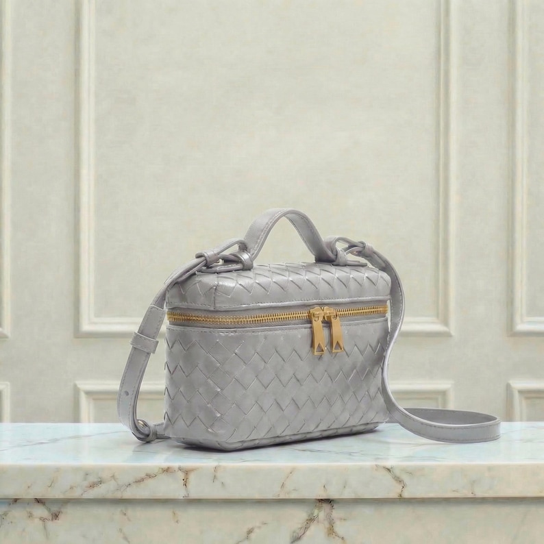 May include: A gray, woven leather crossbody bag with a top handle and a gold-toned zipper. The bag has a rectangular shape and a detachable shoulder strap. The bag is sitting on a marble surface.