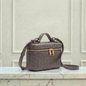 May include: A small, rectangular, woven leather handbag in a muted gray-brown hue. The bag features a top handle, a detachable shoulder strap, and a gold-toned zipper closure. The bag is sitting on a marble surface.
