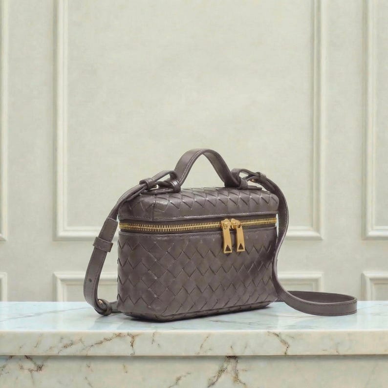 May include: A small, rectangular, woven leather handbag in a muted gray-brown hue. The bag features a top handle, a detachable shoulder strap, and a gold-toned zipper closure. The bag is sitting on a marble surface.