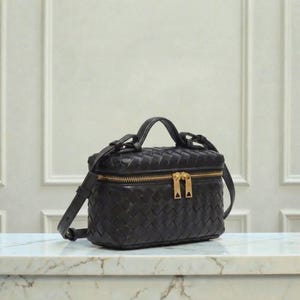 May include: Black leather crossbody bag with a woven design. The bag features a top handle, a detachable shoulder strap, and a gold-tone zipper closure. The bag is sitting on a marble surface.