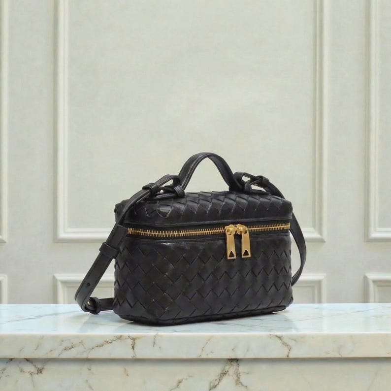 May include: Black leather crossbody bag with a woven design. The bag features a top handle, a detachable shoulder strap, and a gold-tone zipper closure. The bag is sitting on a marble surface.