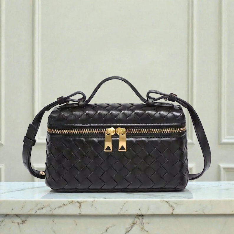 May include: Black woven leather crossbody bag with a gold zipper and handle. The bag has a rectangular shape with a top handle and a long, adjustable strap. The zipper has two gold pulls. The bag is sitting on a marble surface.