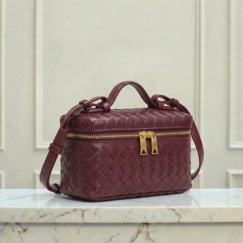 May include: A burgundy leather vanity case with a woven pattern. The bag has a top handle, a detachable shoulder strap, and a gold-tone zipper. The bag is sitting on a marble surface.