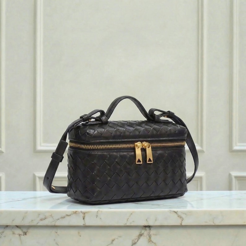 May include: Black leather handbag with a woven design. The bag has a rectangular shape with a top handle and a detachable shoulder strap. It features a gold-colored zipper and hardware.
