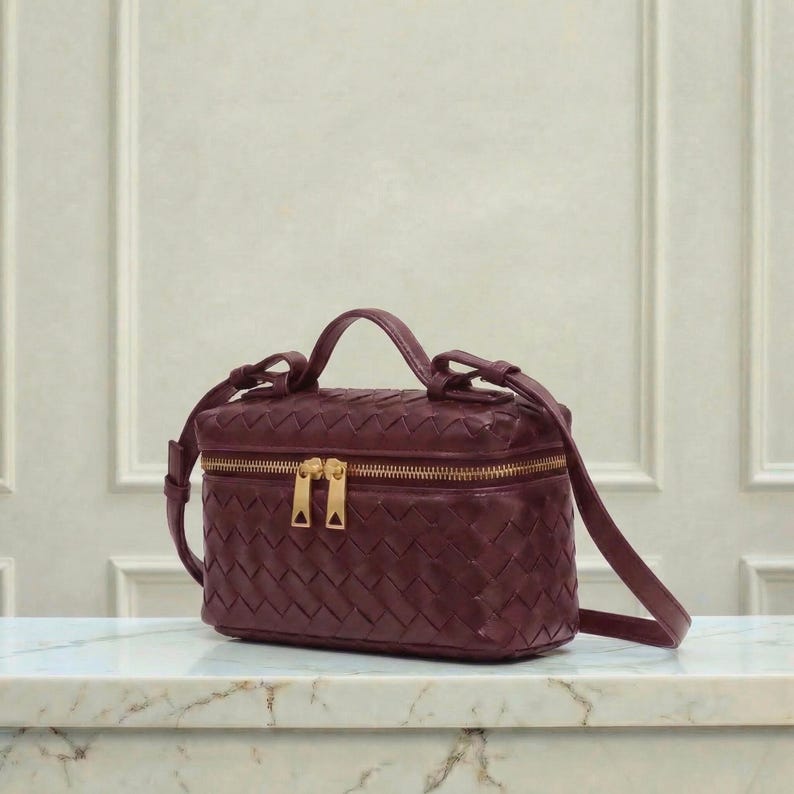 May include: A burgundy leather handbag with a woven pattern. The bag has a rectangular shape with a top handle and a gold-colored zipper. It also features a detachable shoulder strap.