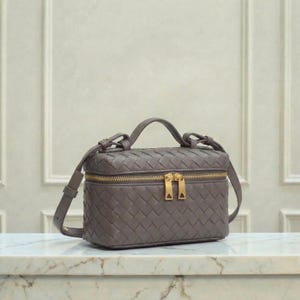 May include: A gray, rectangular handbag with a woven pattern. The bag has a top handle and a detachable shoulder strap. It features a gold zipper and hardware. The bag is sitting on a marble surface.