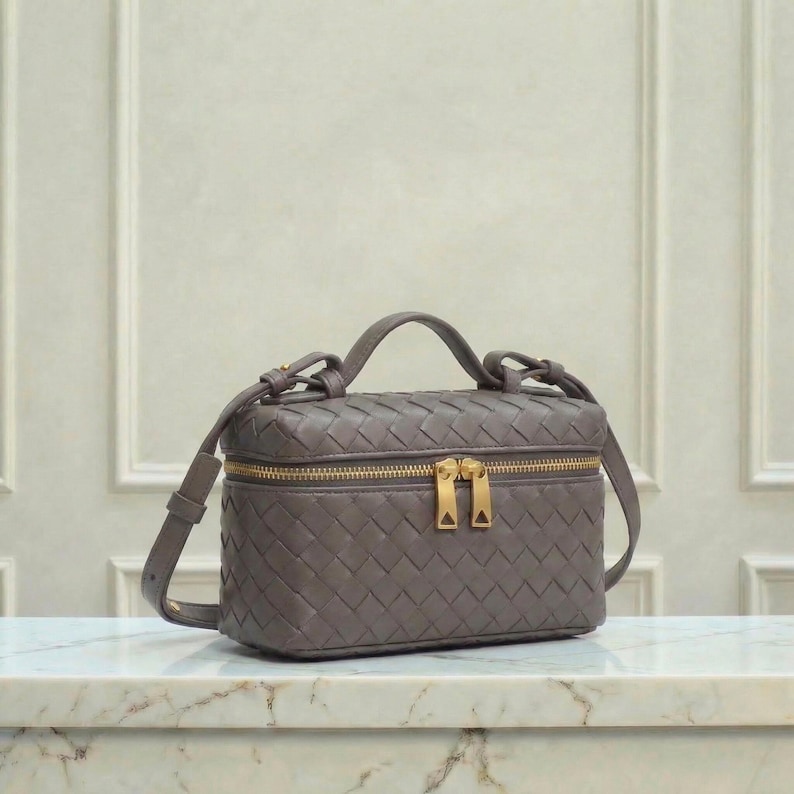 May include: A gray, rectangular handbag with a woven pattern. The bag has a top handle and a detachable shoulder strap. It features a gold zipper and hardware. The bag is sitting on a marble surface.