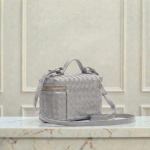 May include: A light gray woven leather crossbody bag with a top handle and a long, adjustable strap. The bag features a gold-toned zipper and hardware. The bag is sitting on a marble surface.