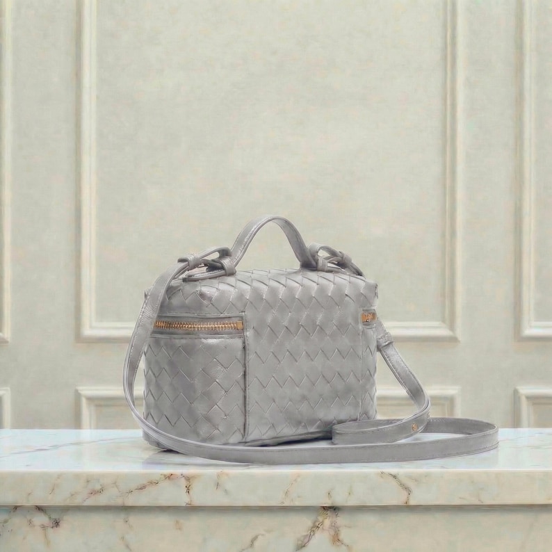 May include: A light gray woven leather crossbody bag with a top handle and a long, adjustable strap. The bag features a gold-toned zipper and hardware. The bag is sitting on a marble surface.